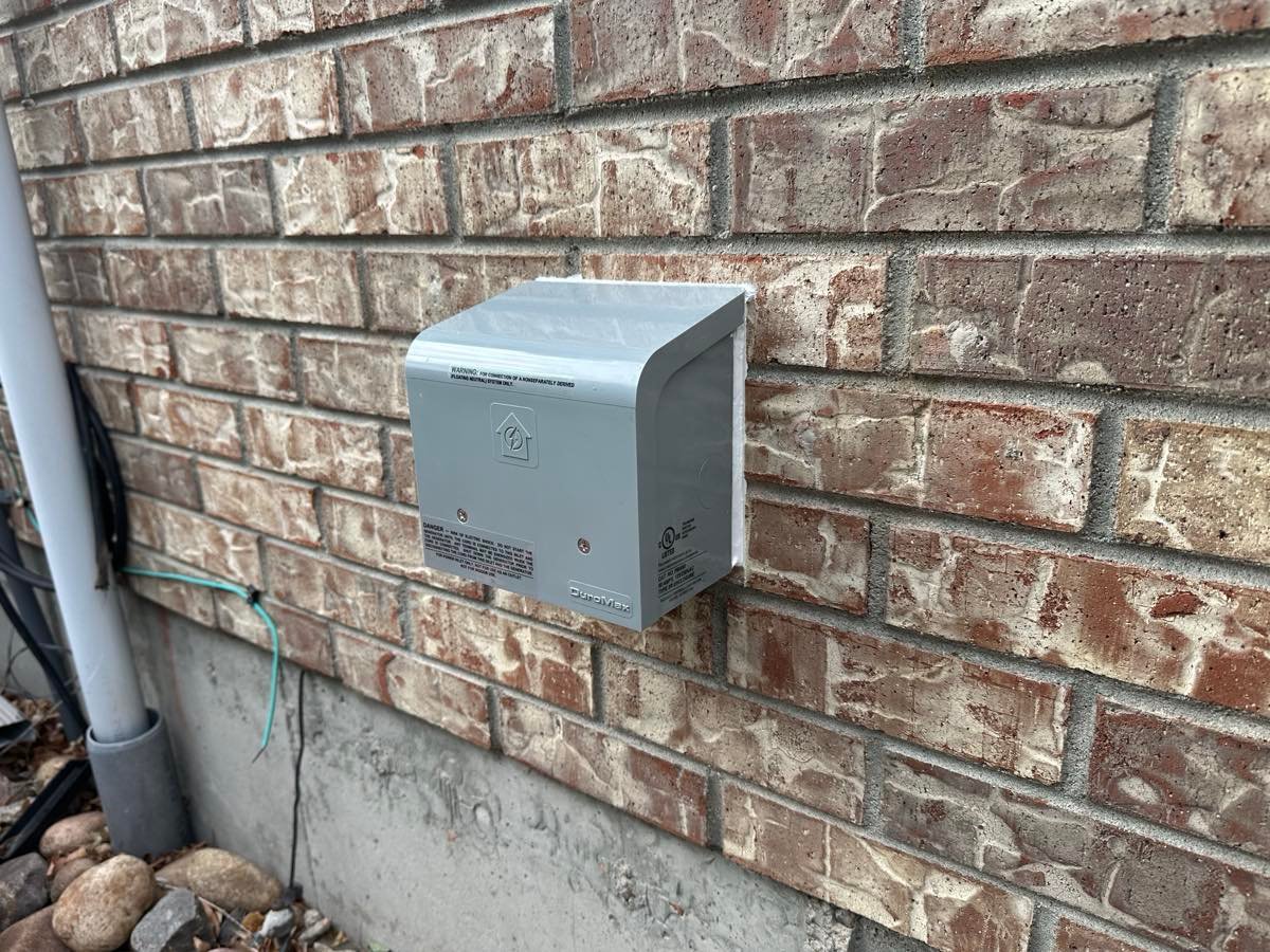 Grey weatherproof generator inlet box mounted on an exterior brick wall