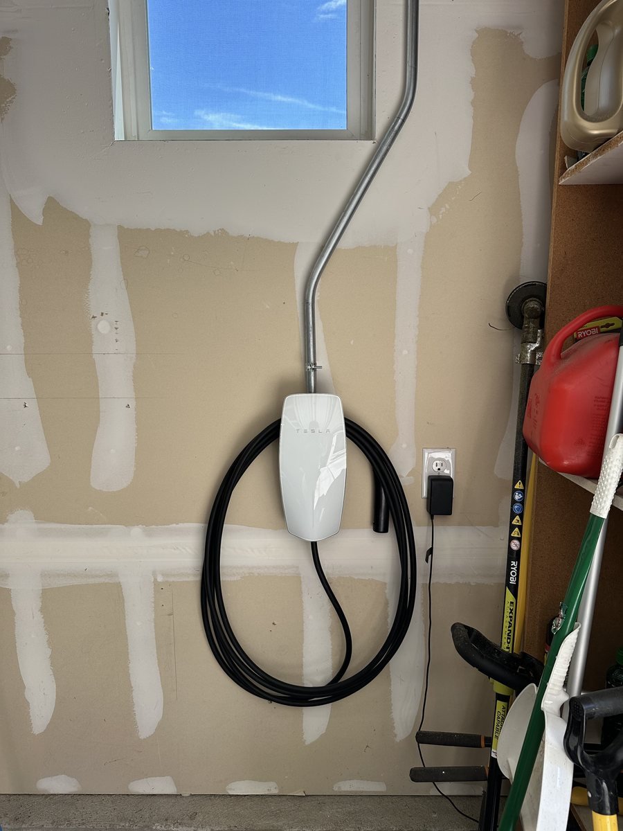 Tesla Wall Connector on a garage wall with conduit bent around a window above it and the cable coiled on its hook